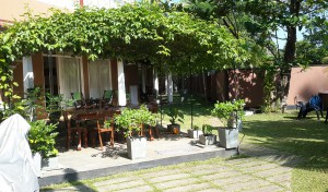 En bild på en pergola med bord och stolar under. I bakgrunden pensionat och trädgård.